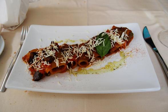 Farbfoto, das einen rechteckigen, leicht kurvigen weißen Teller mit einem Rigatoni-Gericht in Tomatensoße zeigt. Mit einem Basilikumblatt, etwas geriebenem Käse und einer Oliverölspur dekoriert.