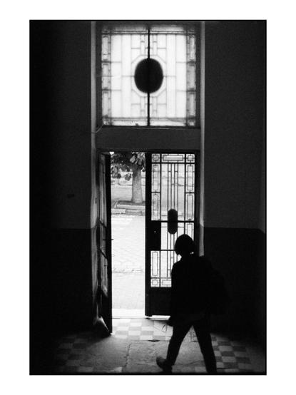 The image is a black and white photograph depicting a person walking through an open doorway. The doorway is framed by a window with a circular design at the top, allowing light to filter into the dimly lit interior space. The floor is tiled with a checkered pattern, and the walls are plain, contributing to the stark contrast between the interior and the brighter exterior. The person, seen from behind, is wearing a backpack and is in motion, suggesting movement from the interior to the exterior. The outside is visible through the doorway, showing a tree and a paved area, indicating a transition from an indoor to an outdoor environment. The overall composition emphasizes the interplay of light and shadow, creating a dramatic and contemplative atmosphere.