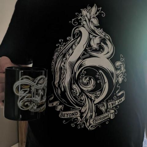 Eine Person trägt ein schwarzes T-Shirt mit kunstvollem, weißem Ornamentdesign in Form eines Violinschlüssels. Der Text lautet „Beyond Tellerrand – Vic Lee“. Sie hält eine schwarze Tasse mit passender Verzierung.