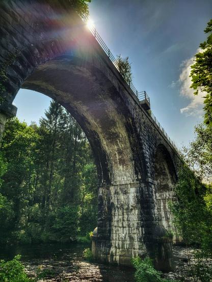 A low-angle shot looking up at a grand, old stone bridge spanning across a dark river. The bridge is made of large, weathered grey stones, with a single, wide arch. Sunlight bursts from behind the top of the bridge, creating a strong lens flare. Lush green trees line both sides of the riverbank, and their reflections are visible in the water. The sky above is bright blue with a few scattered white clouds.