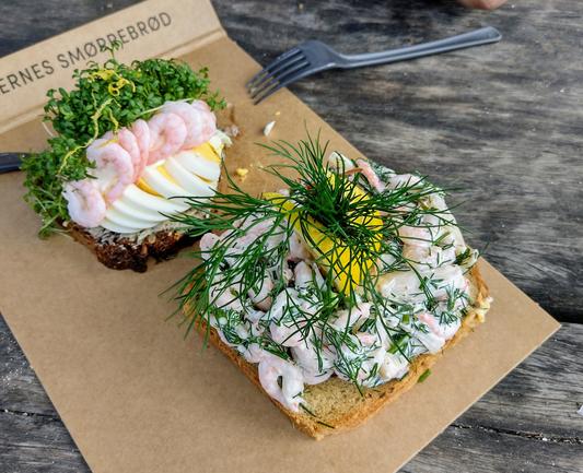 Zwei Smørrebrød (wörtlich übersetzt Butterbrote) auf einer rechteckigen Pappunterlage.

Die Brote liegen leicht versetzt hintereinander. Vorne besteht es aus hellem Brot, das einige Zentimeter dick mit Krabben in einer weißen Sauce mit Kräutern belegt ist. Oben drauf zwei dünne Spalten Zitrone zwischen denen ein dickes Büschel Dill wie ein Blumenstrauß herausragt. 

Schräg einer ein sehr dunkles Brot. Darauf ein komplettes, ein Scheiben geschnittenes Ei. Darauf auf einer Hälfte dick Kresse, auf der anderen Hälfte ca. 10 Garnelen in einer Reihe. Was genau unter der Kresse ist, kann man nicht erkennen. 

Das hintere Brot ist angeschnitten, eine kleine Ecke fehlt, eine Gabel liegt daneben