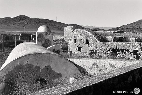 This black and white photo, captured on a Nikon F using Ilford Delta 100 film, showcases abandoned water tanks and cisterns, which supplied water to the Cortijo del Fraile, with rounded roofs crumbling amid tall grass. Hills rise in the background under a clear sky, evoking a timeless scene.