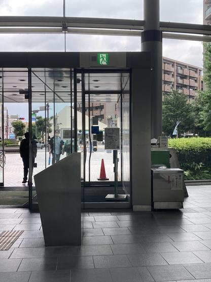 A glass entrance to a building with an emergency exit sign above. Visible outside are pedestrians, a traffic cone, and greenery. An angular metal structure is in the foreground, with a bin beside it. The atmosphere is urban and slightly cloudy.