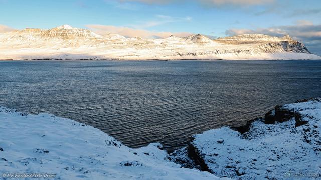 A colour photo of a body of water with sunlit snow-covered mountains on the horizon. The foreground is icy black rocks and, in the centre of the shot, a V-shaped cleft. The sky is fairly clear and the water of the fjord is ruffled.
