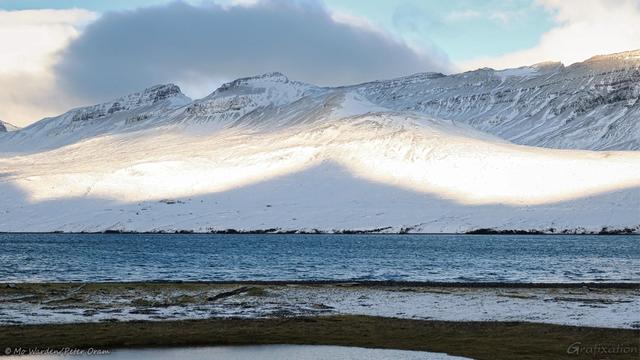 Another colour shot of the same fjord further along the road. The foreground is a dark sandy bank with a sprinkling of snow. On the opposite shore is a snowy mountainside, the sky above it is more cloudy. The peaks of the mountains and the water below are in shadow but the flanks are sunlit. In the centre is the pointed shadow of the mountain behind us, cast across the snowfield.