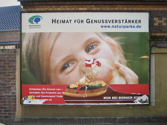 das bild im querformat zeigt eine große werbetafel an einer hauswand. das plakat wirbt mit dem slogan "heimat für genussverstärker" für "naturparke deutschland".
mittig und großflächig ist der kopf eines kindes, welches in einen apfel beißt. das plakat ist beschädigt und wurde teilweise abgerissen. auf höhe des mundes des kindes tritt ein teil eines burgers einer großen burgerkette zum vorschein und erweckt den eindruck, das kind würde den burger essen.