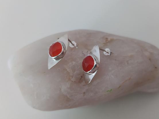 a pair of silver romboid stud earrings with an orange gemstone each. they are placed on a light gray pebble
