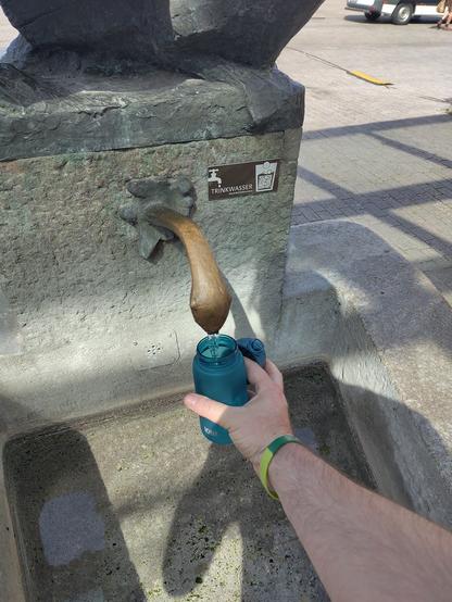 Eine Hand hält eine Trinkflasche und füllt sie mit Wasser aus dem Marktbärbel-Brunnen am Unteren Markt in Würzburg.