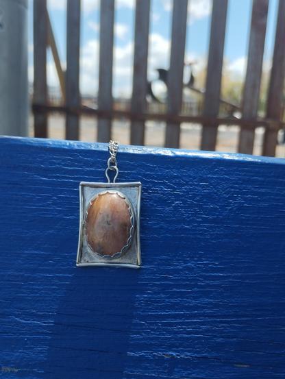 a pendant, it has an oval stone in silver rectangular frame. It's placed on a blue painted wood