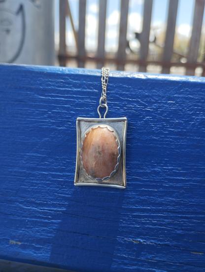a pendant, it has an oval stone in silver rectangular frame. It's placed on a blue painted wood
