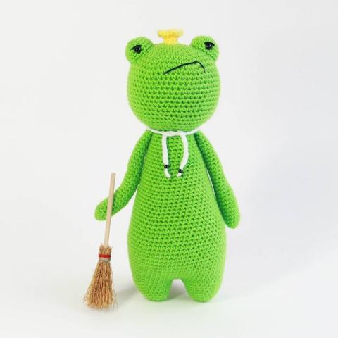 Picture of a crocheted amigurumi frog with a crown and cape standing upright, with a moody face, holding a broom.