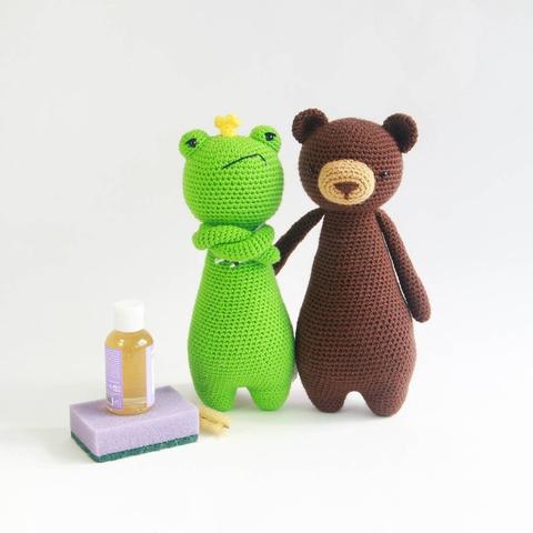 Picture of a crocheted amigurumi frog and bear standing upright. The frog is looking moody. In front of them are a few cleaning supplies.
