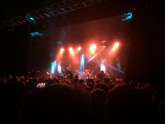 A photograph taken from the side of the O2 Academy in Bristol, UK showing the Devin Townsend Project in the middle of a performance. The stage is primarily lit by six downward-pointing orange spotlights. A half-dozen fans of pale blue light play upwards and downwards, illuminating sections of the lighting truss at the top of the shot. In the audience, a single mobile phone screen is visible as someone records the song. 

Dev is front and centre, of course, holding a guitar and singing. The rest of the band (Dave on guitar, Ryan on drums and Brian on bass) have their heads down, thundering through a particularly heavy section.
