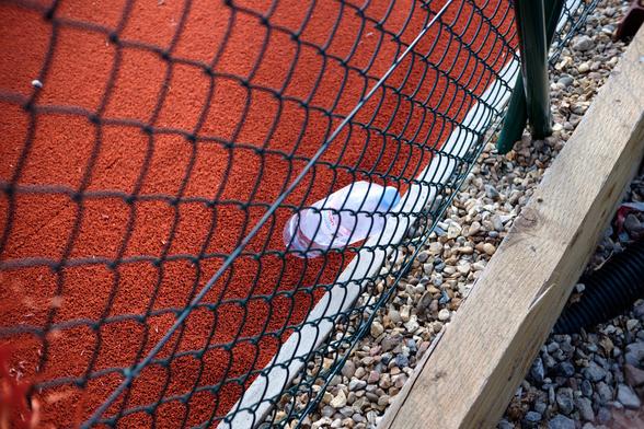 A photo from behind a fence of a tennis court. A bottle of water takes the centre of the frame. The rest of the frame consists of diagonal layers: red court floor, net wire fence, white border, rubble cover and wooden board