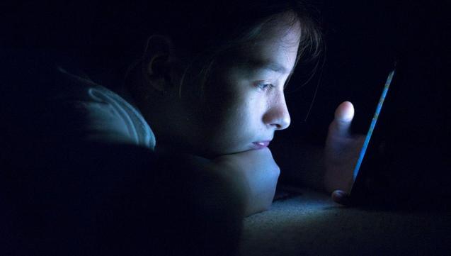 Un enfant regarde l'écran d'un smartphone, dans le noir, éclairé seulement par celui-ci.
