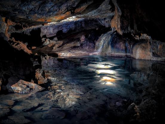 The interior of the Erdmannshöhle cave, dominated by a large, still body of water reflecting light from above. The cave walls and ceiling are rugged and irregular, showcasing various rock formations in shades of dark brown, orange, and hints of blue and purple. Along the edges of the water and on the cave walls, there are visible stalactites and stalagmites.