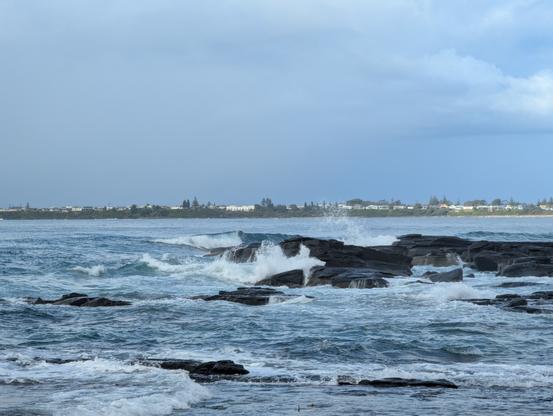 Ocean rocks hammered by incoming waves with a suburban headland in the back
