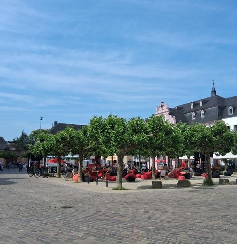 Unter vielen Platanen sind Sitzmöglichkeiten aufgestellt, alle in beiger, schwarzer oder rot-orangener Farbe. Der Platz vor dem Dom wird eingegrenzt von quaderförmigen Steinen.