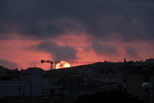 Picture of the sun setting behind a hill in Amadora. The sun is partially covered by clouds and is also being framed by the silhouette of a construction crane.