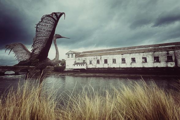 A dramatic, wide shot under a brooding, cloudy sky features a large, dark metal heron sculpture in the foreground on the left, its wings spread. In the midground, a dark river flows, reflecting the sky. On the right bank of the river stands a long, old, white brick building with many windows, suggesting an industrial past. Tall, golden-brown grasses are blurred in the immediate foreground, adding depth to the scene. A small stone bridge is visible in the far left background.