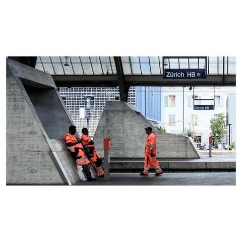 Sene at Zurich Main station, there are shields labeled "Zürich HB." Three workers wearing bright orange safety vests and reflective stripes are present in the scene. Two of the workers are seated on a concrete structure, while the third worker is walking nearby. The orange stands out against the grey concrete background. (with help of Mistral.ai)