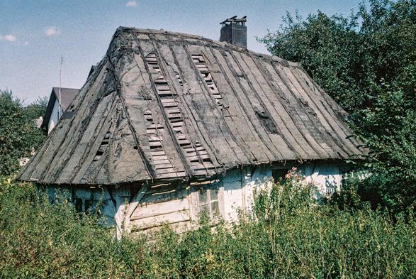 The image shows an old, dilapidated wooden house surrounded by overgrown vegetation. The roof is severely damaged, with large patches of torn and missing covering, revealing the wooden slats beneath. The structure appears weathered and neglected, with peeling white paint on the walls and broken windows. A metal chimney protrudes from the roof, suggesting the house was once inhabited. In the background, modern houses with intact roofs are visible, creating a contrast between abandonment and contemporary life. The scene evokes a sense of decay, memory, and the passage of time.