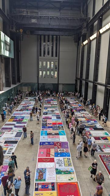 Many colourful quilt panels laid out on the floor of a large room in three long columns. People stand at the borders, reading the messages on the panels.