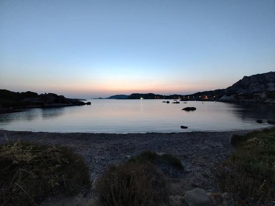 Una spiaggia in una piccola baia, un'ora dopo il tramonto, rosso all'orizzonte, acqua calma, scogli che affiorano, luci sulla costa in lontananza. Sensazione di calma e di pace.