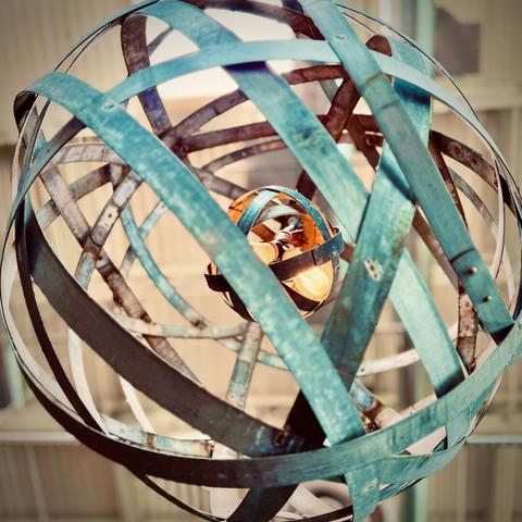A loose, spherical chandelier comprised of turquoise, criss-crossing metal slats wrapped around a much smaller, similar sphere housing two Edison bulbs hangs from a tall, white ceiling in the distance.