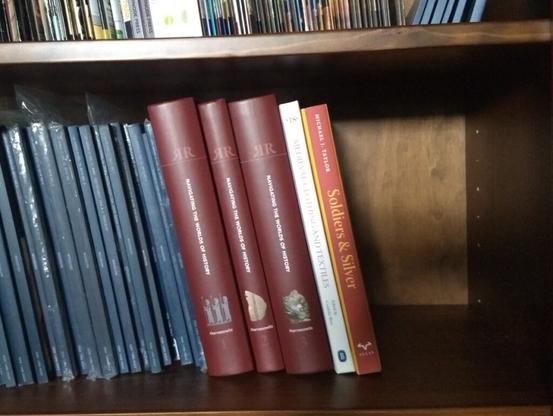 A wooden bookshelf stained dark brown with many issues of the classics journal "Mouseion" in its dark blue softcover binding, three burgundy hardcover volumes of "Festschrift Rollinger", a white hardcover issue of "Medieval Clothing and Textiles," and the red and yellow hardcover "Soldiers and Silver" by Michael J. Taylor sitting on it