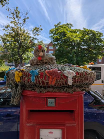 Red postbox on a street in Southport, UK covered on top with a yarn-stitched or crocheted chicken 🐔