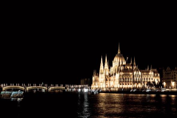 Das Bild zeigt eine nächtliche Stadtansicht, dominiert vom imposanten Parlamentsgebäude in Budapest, dessen goldene Beleuchtung sich im dunklen Flusswasser spiegelt. Statt klarer, scharfer Konturen entsteht durch die Bewegung der Kamera ein leicht verzerrtes, fließendes Lichtspiel, das eine fast malerische Wirkung entfaltet. Besonders die hellen Strukturen der Brücke und die Umrisse der Architektur erscheinen wie sanft verwischte Pinselstriche auf einem dunklen Hintergrund.

Die bewusste Integration von ICM-Technik erzeugt einen spannenden Effekt, bei dem die Stadt weniger statisch wirkt – vielmehr scheint sie in Bewegung zu sein, als würde die Szene durch ein visuelles Echo pulsieren. Das Licht wird zum Hauptakteur, während feste Formen zugunsten eines impressionistischen Eindrucks aufgelöst werden.

Das Bild vermittelt eine Mischung aus Realität und Abstraktion, bei der Architektur, Licht und Wasser ineinanderfließen. Dadurch entsteht eine einzigartige Stimmung, die Budapest nicht nur als Stadt zeigt, sondern als lebendiges, strömendes Kunstwerk.