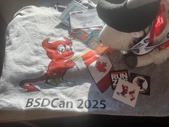 Groff the BSD goat waking up with a selection of the stickers you can grab at the registration desk and the BADCan 2025 t-shirt.