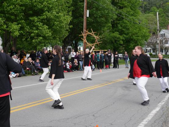 Six longsword dancers with red shirts, black tunics, and white trousers dancing in a street. They are moving in a circle. One is holding eight longswords weaved into a star shape above his head. There is an accordion player partly visible on the left. Behind them an audience including some morris dancers.