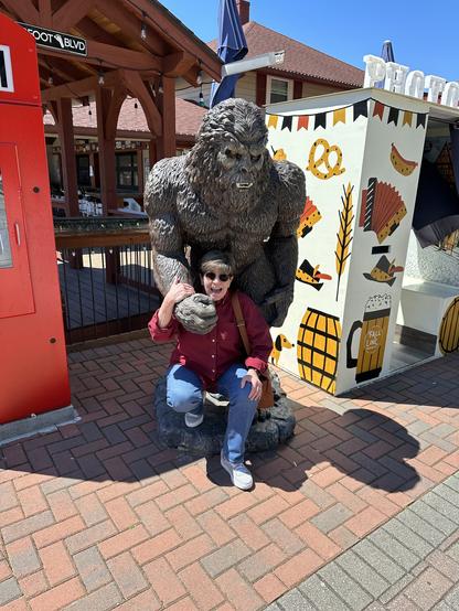 A woman in sunglasses smiles while posing with a Bigfoot statue. The setting is colorful and outdoors, with festive decor and a sunny ambiance.