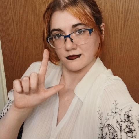 A photo of Goddexx Andi with their hair up, make up done, and holding up a loser symbol while looking at the camera while smirking
