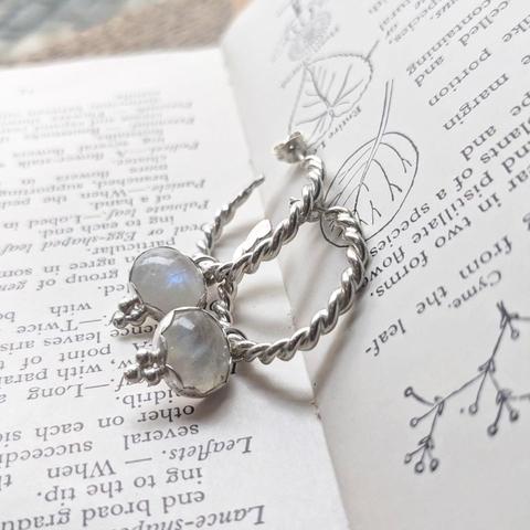 A pair of hoop earrings lying in the fold of a book. The earrings are made of twisted silver wire and are decorated with moonstones.