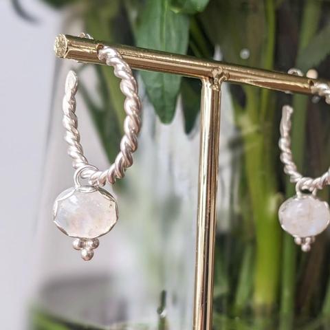 Silver earrings on a brass display stand. The earrings are made of twisted silver wire and are decorated with moonstones. There is a part of a green plant visible in the background.