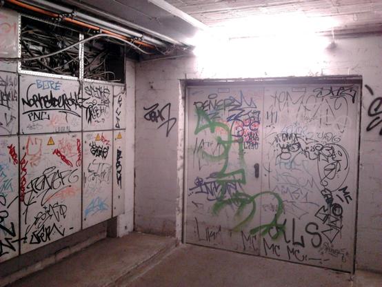 Foto einer hässlichen, dreckigen, mit Graffiti übersäten Tür im Sockelgeschoss der Ruine des Ihmezentrums in Hannover-Linden. An der Seite der Tür ein oben geöffneter elektrischer Schaltkasten und offen verlegte Kabel und Rohre.