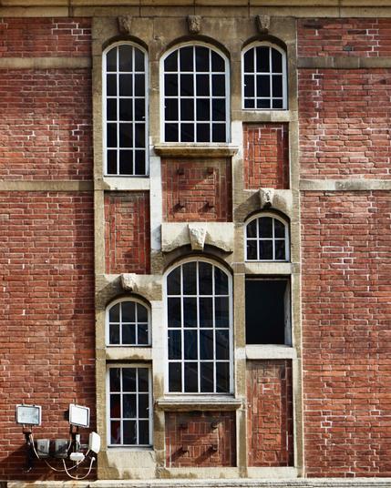 Détail d’une façade en briques du début du XXe siècle. On voit deux séries superposées de petites fenêtres décalées les unes par rapport aux autres car vraisemblablement alignées sur un escalier à l’intérieur et qui sont encadrées de grès.