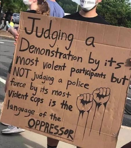 Cardboard sign held by a masked person features text: "Judging a demonstration by its most violent participants but NOT judging a police force by its most violent cops is the language of the oppressor." Two raised fists are drawn on it. Nearby people stand outdoors.