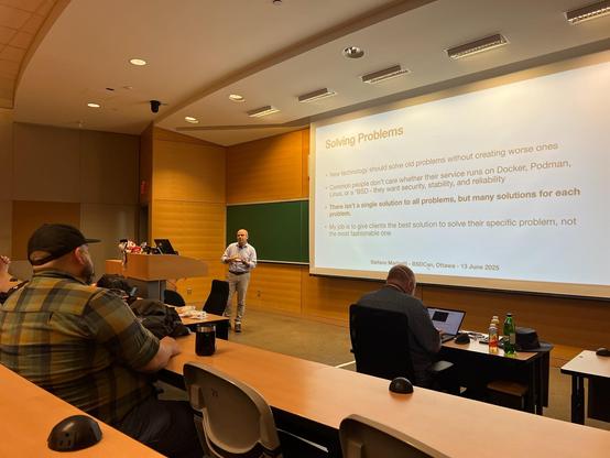 A speaker (me) stands in front of a classroom-style lecture hall giving a presentation - the current slide is titled "Solving Problems" - BSDCan in Ottawa on 13 June 2025. The slide emphasizes practical solutions over trendy technologies, stating that clients value stability and reliability more than whether a service runs on Docker, Podman, Linux, or BSD. Several audience members are seated, some with laptops and drinks, attentively listening. The room has a large projection screen, wooden paneling, and a chalkboard.