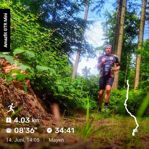Person joggt auf einem von Wald umgebenen Naturweg. Sie trägt ein schwarzes Sportoutfit mit bunten Mustern. Links liegt ein umgestürzter Baumstamm, rechts ist dichter grüner Bewuchs. Auf dem Bild eingeblendet sind Trainingsdaten: 4,03 km in 34:41 Minuten mit einer Pace von 08:36 min/km. Unten rechts eine Karte der gelaufenen Strecke, oben Datum (14. Juni), Uhrzeit (14:05 Uhr) und Ort (Mayen). Links steht "Amazfit GTR Mini"