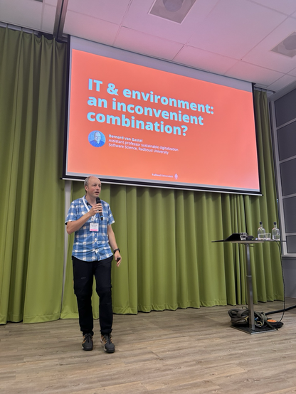 Bernard van Gastel op het podium links op de foto. Achte them een heel groot scherm met een oranje vlak en de tekst “IT and environment an inconvenient combination?
