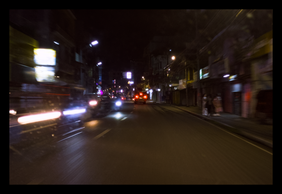 This is a motion-blurred, long-exposure photograph of a city street at night, captured from the perspective of a moving vehicle. The foreground and sides are streaked with horizontal lines of light: vibrant white and blue from oncoming headlights on the left, and the red glow of taillights from cars ahead. The road itself is a dark, reflective surface, catching the gleam of various light sources. Lining the street are multi-story buildings, their details obscured by the motion. Some storefronts on the right appear closed with shutters down, while illuminated signs and streetlights cast a mix of warm yellow and cool white light onto the scene. On the right sidewalk, the faint, ghostly figures of a few people can be seen, also blurred by the camera's movement.