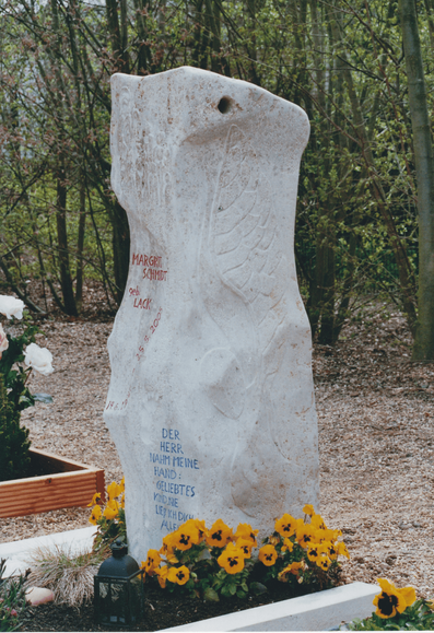 Schrägansicht von rechts auf die helle Stele. Oben durchzieht ein faustgroßes Loch die Spitze. Rechtsflanke trägt ein fein eingemeißeltes, nach unten schwingendes Flügelrelief; darunter ein glatter runder Teller wie Sonne / Herz. Die Vorderseite biegt zur Kamera und zeigt rote Inschrift. Am Fuß halbkreisförmig leuchtend gelbe Stiefmütterchen mit schwarzer Zeichnung. Links davor eine schwarze Metalllaterne, daneben ein rosafarbenes Steinherz und ein Büschel hell-grüner Ziergräser. Die Grabfläche bleibt zweigeteilt: rechts dunkle Erde, links – am unteren Bildrand sichtbar – der geschwungene Streifen aus schneeweißen Kieseln. Steinrahmen aus hellem Marmor fasst das Grab. Links ragt das Nachbargrab mit Rosen in Holzumrandung ins Bild. Hintergrund: dichter, grüner Busch- und Jungwald. Das Gesamtbild wirkt hell, organisch und tröstlich – der Engelsflügel scheint die Inschrift schützend zu umfangen.