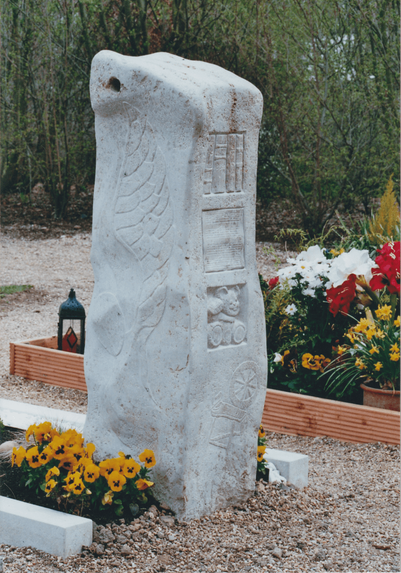 Rückansicht der Stele. Links ragt der große, fein gefiederte Engelsflügel der Seitenfläche ins Bild.
Die Rückseite ist in vier quadratische Relief­felder übereinander gegliedert: Bücherregal – fünf aufrechte Buchrücken. Notenblatt – rechteckiges Feld mit dicht gravierten Noten­linien; ein undeutliches Oval markiert vielleicht den Notenschlüssel, Hinweis auf musikalische Leidenschaft. Teddybär – runder Kopf mit Knopfnase, steht. Spinnrad – großes Rad mit sechs Speichen, Ständer und Trittbrett, Sinnbild für Handwerk und Lebensfaden.
Flügel, Noten, Bücher, Spielzeug und Spinnrad bilden eine vertikale Lebens­geschichte, geschützt vom Engelsflügel. Am Fuß halbkreisförmig leuchtend gelbe Stiefmütterchen, links davor eine schwarze Laterne, rechts bunte Grabsträuße in Holzumrandung. Das Ensemble wirkt verspielt und zugleich würdevoll.