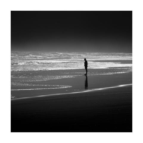 A square black-and-white photograph, framed with a white border, showing a solitary person standing at the edge of the sea. The figure is captured in profile, silhouetted against the bright, shimmering surface of the wet sand and distant waves. Light reflections form a horizontal band across the midsection of the image, separating the dark beach in the foreground from the misty, glowing ocean in the distance. The person’s faint reflection is visible beneath their feet. The overall atmosphere is quiet and minimal, with strong tonal contrast.