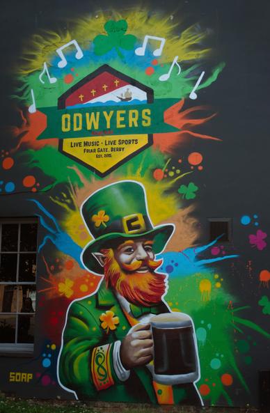 Artwork on the end of Odwyers Irish Pub in Derby, a black wall with a ginger haired leprechaun wearing green holding stout