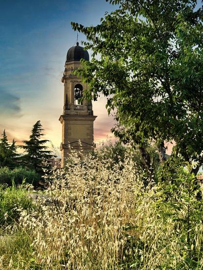 Ein Glockenturm fotografiert bei Sonnenuntergang zwischen hohen Gräsern und Bäumen. Der Himmel weist sanfte Pastelltöne auf und verleiht der Szene eine heitere Atmosphäre.